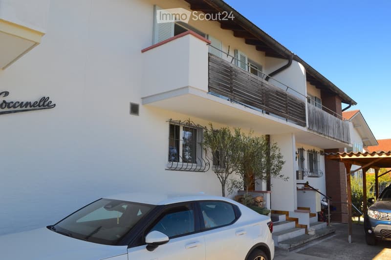 Two-story white building with a balcony, small garden, and parked cars in front.