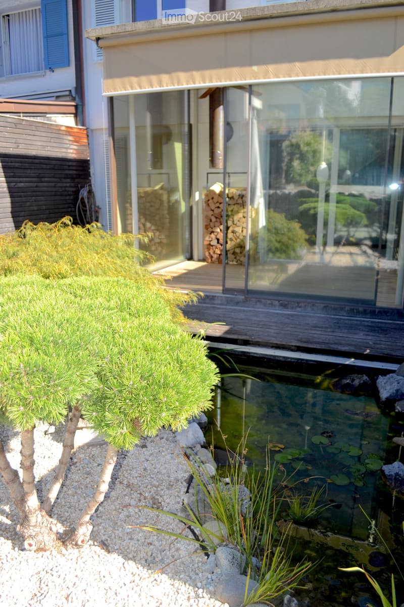 Modern house with a glass extension, a small pond with lily pads, a small tree, and a stack of firewood near the entrance.