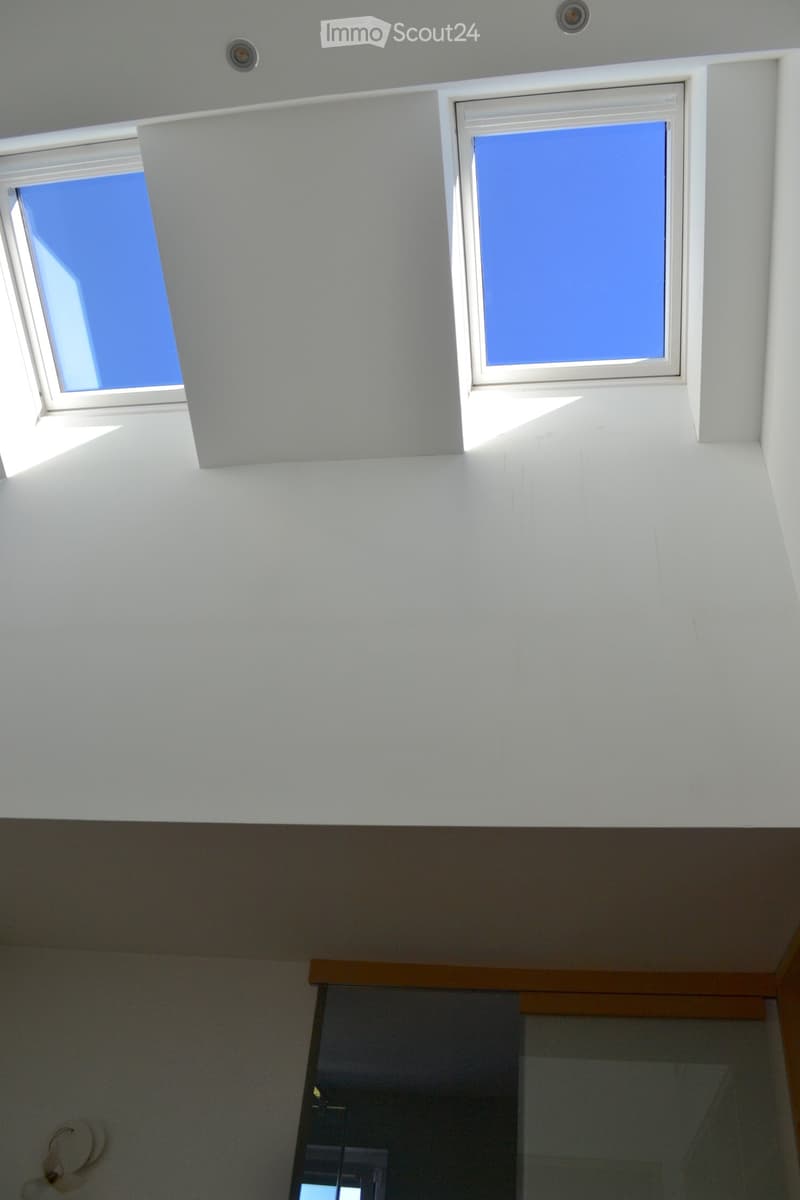 Bright, modern corridor with white walls and ceiling, featuring two skylights and a glass door leading to another room.