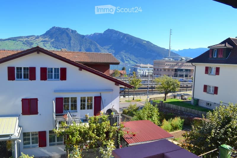 Two-story house with red shutters and a tiled roof, situated in a scenic area with mountains in the background and a view of a lake and town.