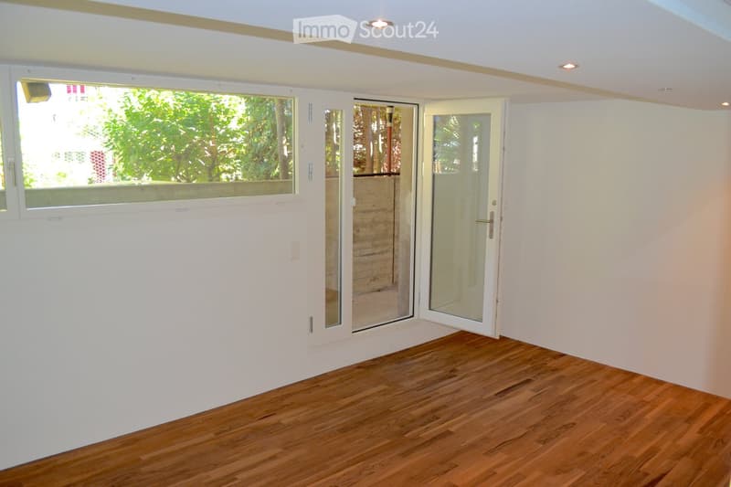 Empty room with wooden flooring, white walls, and large windows with sliding glass doors leading to an outdoor area.