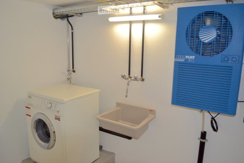 A compact bathroom with a white washing machine, a beige sink, a faucet, and a blue fan unit mounted on the wall.