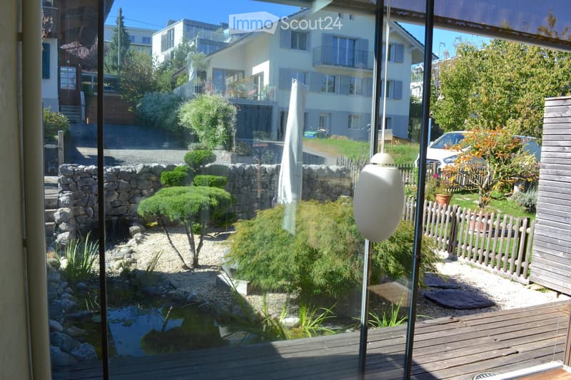 View from inside a house looking out onto a well-maintained garden with a small pond, stone walls, neatly trimmed shrubs, and a wooden deck.