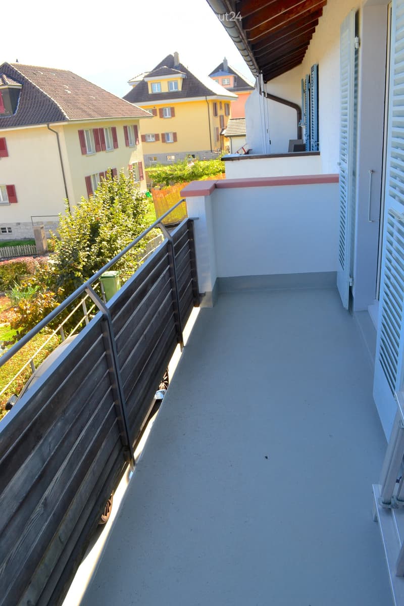 Balcony with wooden railing, concrete floor, and view of neighboring houses and greenery.