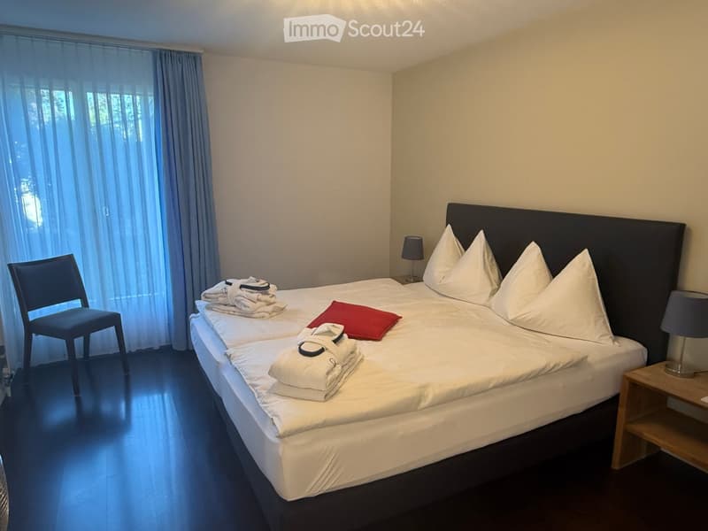 A well-lit bedroom with a double bed, two nightstands, a chair, and neatly arranged towels and pillows on the bed.