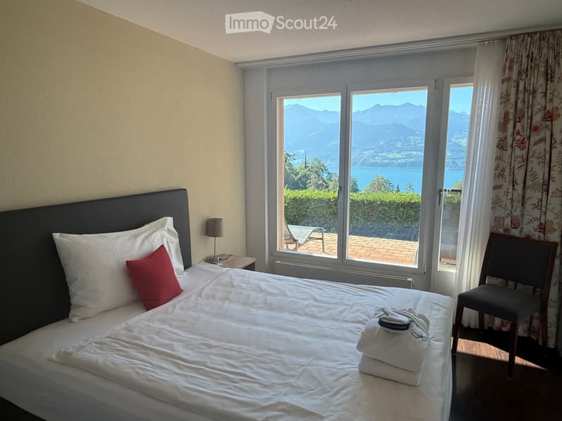 Cozy bedroom with a large bed, white linens, red accent pillow, nightstand with lamp, and a scenic mountain view through large windows.