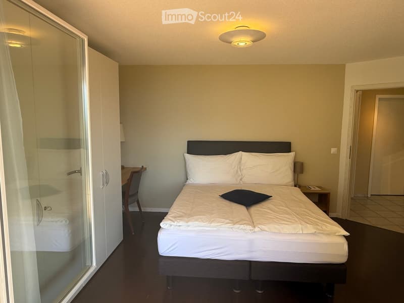 Modern bedroom with a double bed, nightstand, chair, and wardrobe. The room features a sleek design with neutral tones and contemporary furniture.