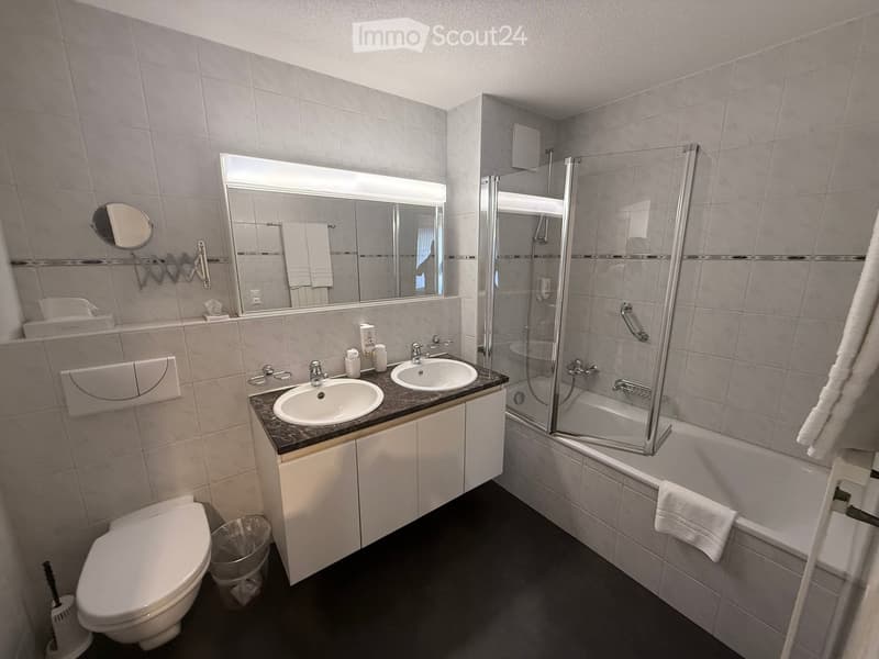 Modern bathroom with double sink, toilet, bathtub with glass shower enclosure, and mirrored cabinet.