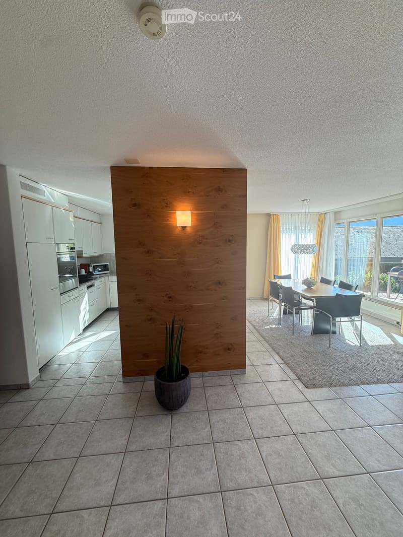 Open-plan living and dining area with tiled flooring, white kitchen cabinets, and a wooden partition separating the spaces. The dining area features a wooden table with chairs and a chandelier hanging above.
