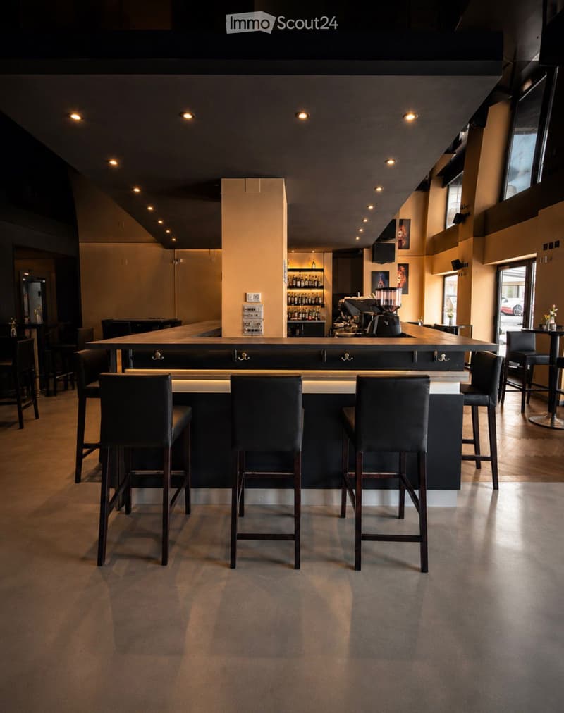 Modern bar area with a long counter, several high stools, a coffee machine, shelves with bottles, and additional seating area with tables and chairs.