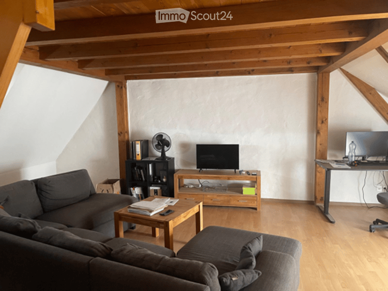 Living room with wooden beams, grey sofa, coffee table, TV stand, fan, bookshelf, and a small table with a chair.