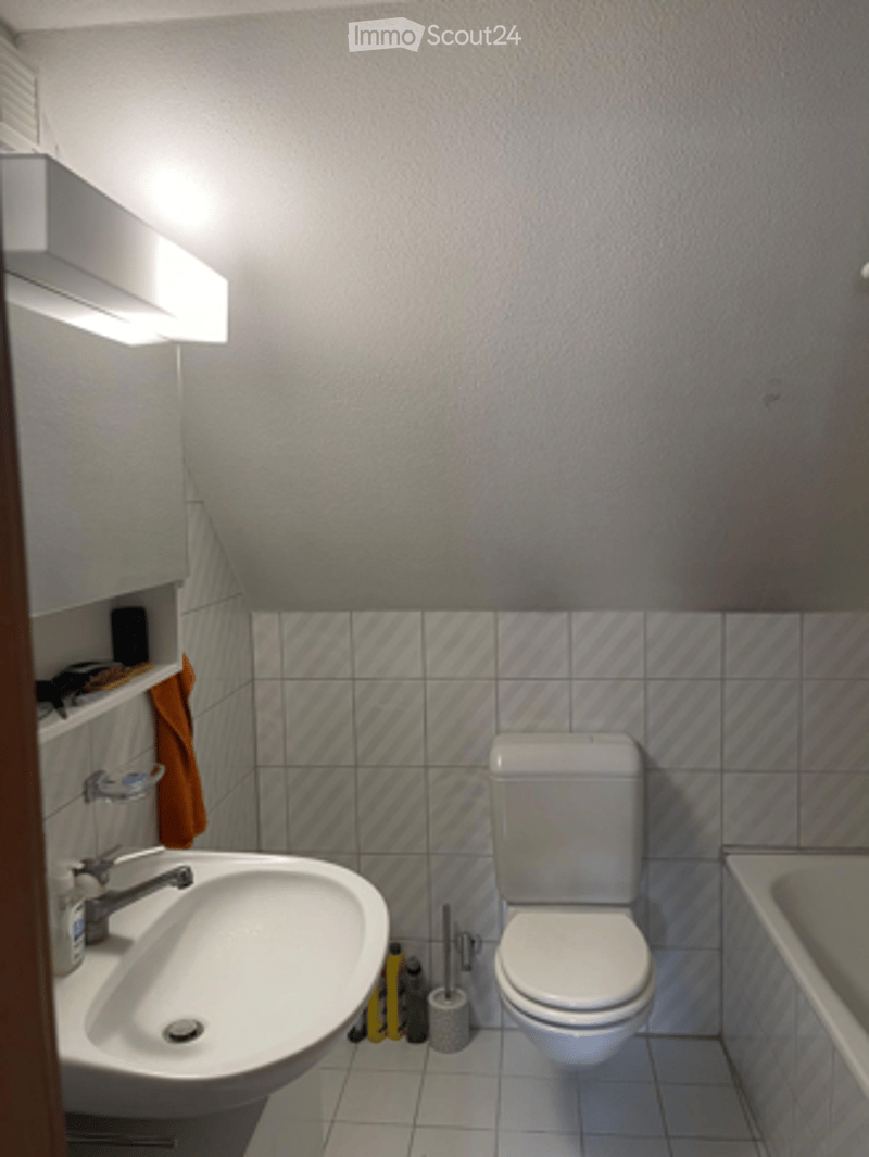 Modern bathroom with white tiles, a sink, toilet, bathtub, and a shelf with a towel and toiletries.