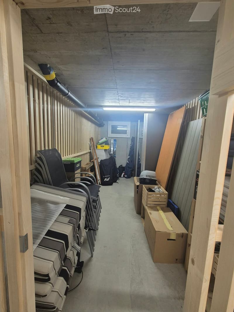 A narrow storage room with wooden walls and a concrete floor, containing stacked chairs, backpacks, snowboards, and boxes.