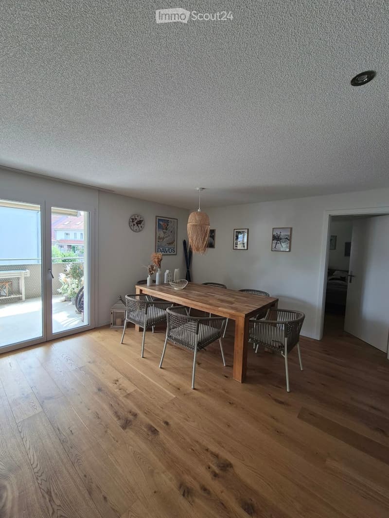 Dining room with wooden floor, large wooden dining table, four chairs, decorative vases, picture frames, and a wicker hanging lamp.