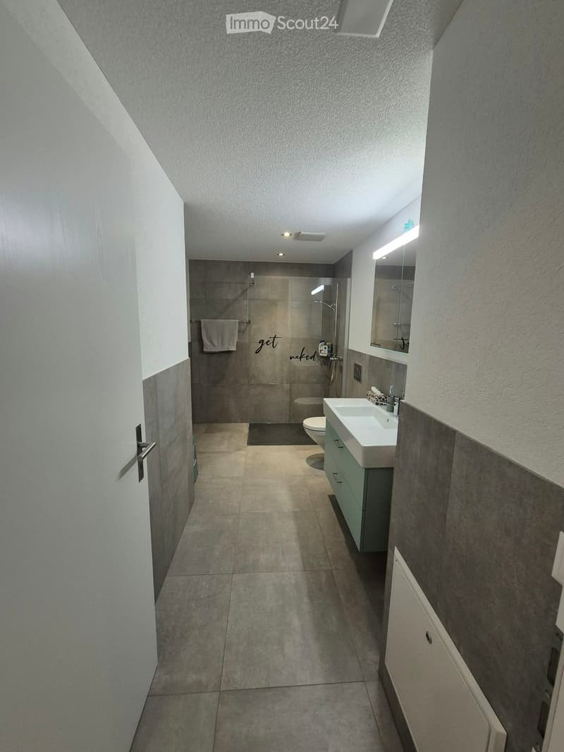 Modern bathroom with a sink, mirror, toilet, and shower area. The space features light-colored tiles and a sleek design.