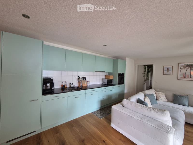 A modern, open-plan living and kitchen area with light blue cabinets, a white sofa, and a coffee table. The kitchen features a sink, oven, microwave, and coffee machine.