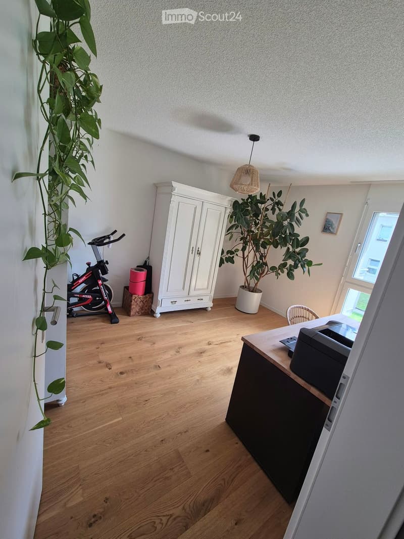 A well-lit room with wooden flooring, a white cabinet, an exercise bike, a desk with a printer, and potted plants.