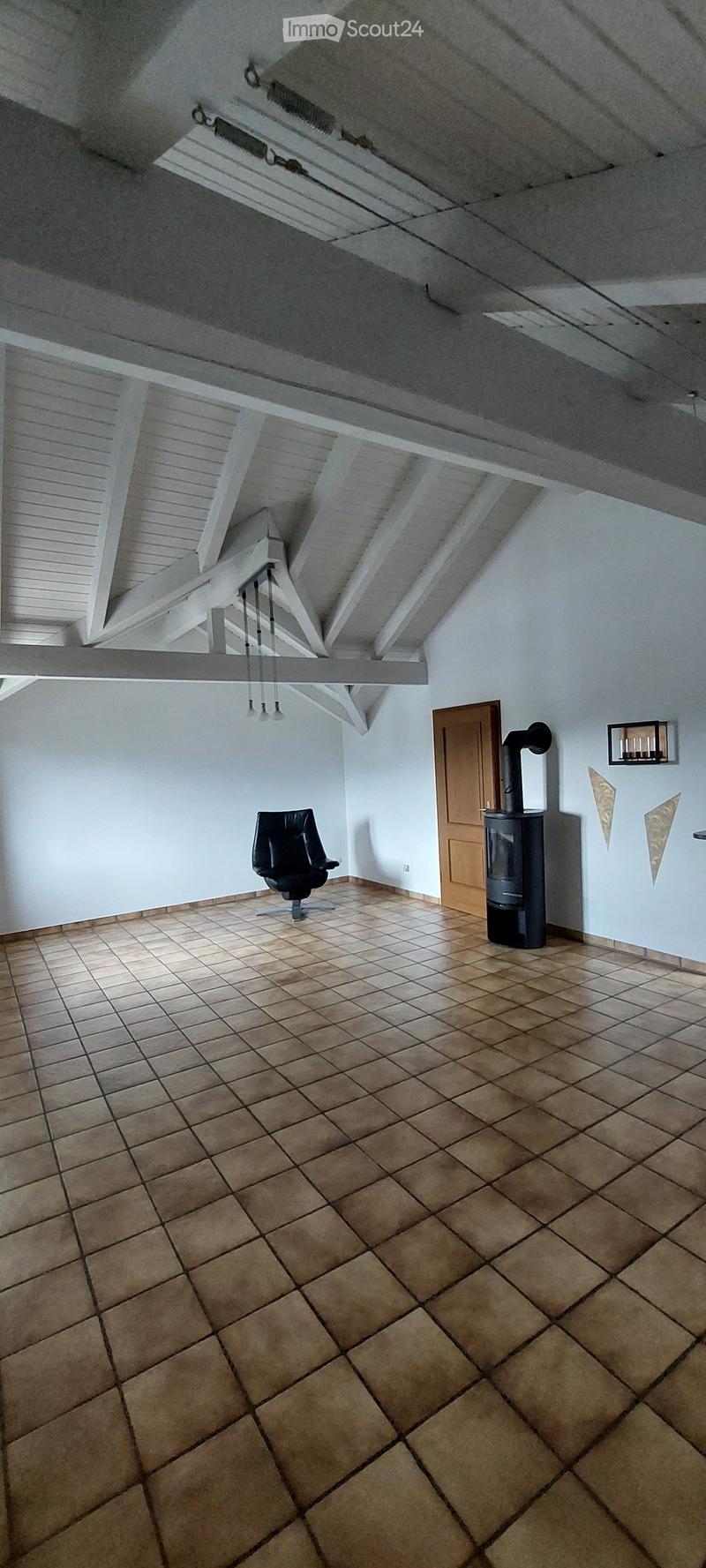 Room with exposed wooden beams, tiled floor, a single black chair, a wooden door, and a wood stove.