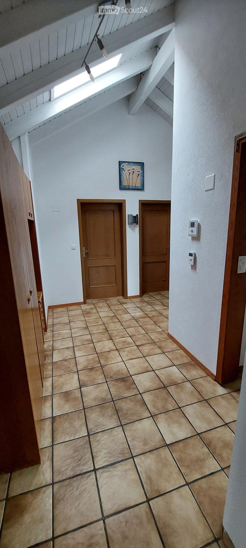 A hallway with tiled flooring, white walls, and two wooden doors. A small window is visible at the top of the wall, and a framed picture hangs on the wall.