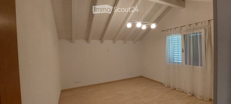 Empty room with wooden flooring, white walls, and a slanted wooden ceiling. A window with white blinds and sheer curtains is visible, along with a modern pendant light fixture.