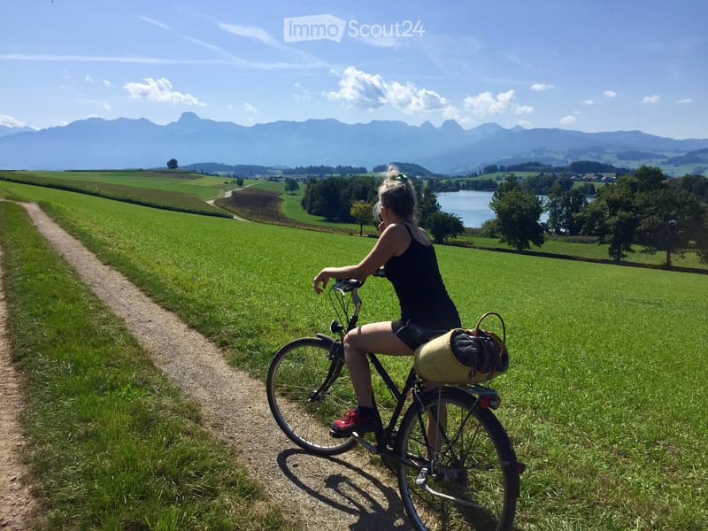 A woman rides a bicycle on a dirt path through a lush green field with a scenic mountain backdrop and a lake in the distance.