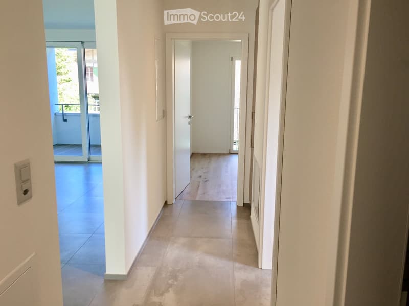 Empty hallway with white walls, tiled floor, and a door leading to a balcony area with wooden flooring.