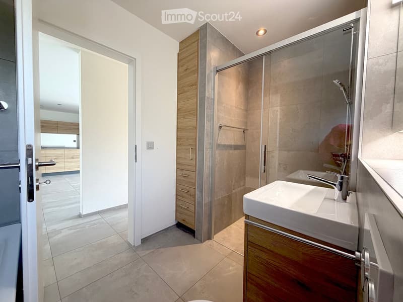 Modern bathroom with a white sink, wooden cabinet, glass shower stall, and a visible kitchen area through an open doorway.