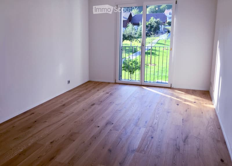 Empty room with light wooden flooring and white walls, featuring large glass doors leading to a green outdoor area.