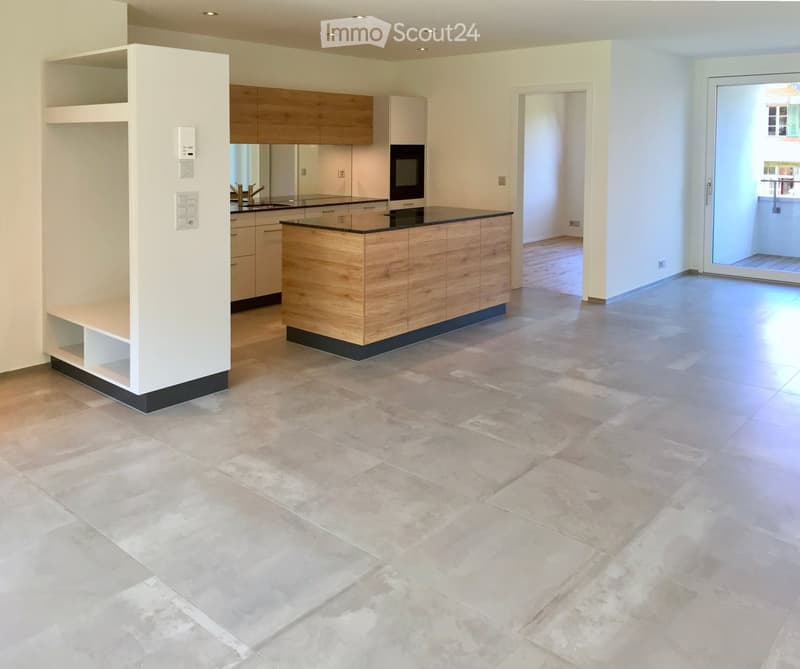 Modern kitchen with light wooden cabinets, a central island, and a large open space. The floor is tiled in a light beige color, and there is a sliding glass door leading to another room.