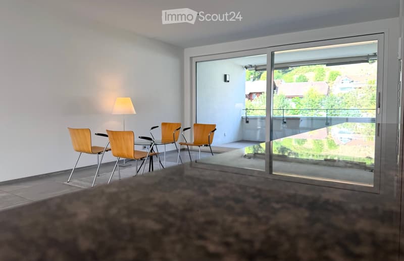 Bright, modern room with large glass sliding doors leading to a balcony, four wooden chairs around a small table, and a floor lamp near the wall.