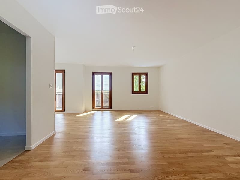 Empty room with light wooden flooring, white walls, and large windows and doors allowing natural light to enter.