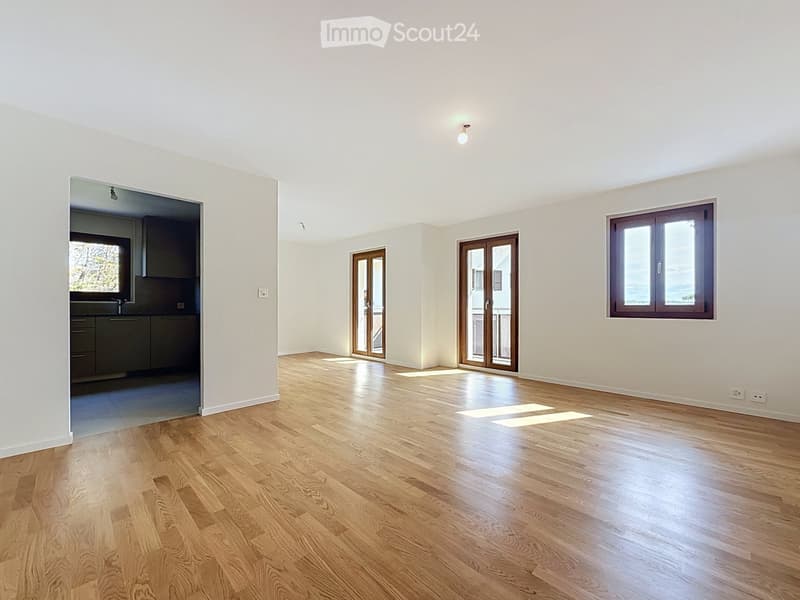 Empty room with light-colored walls, wooden flooring, and large windows. Features include a sliding glass door and an adjacent kitchen area visible through an open doorway.