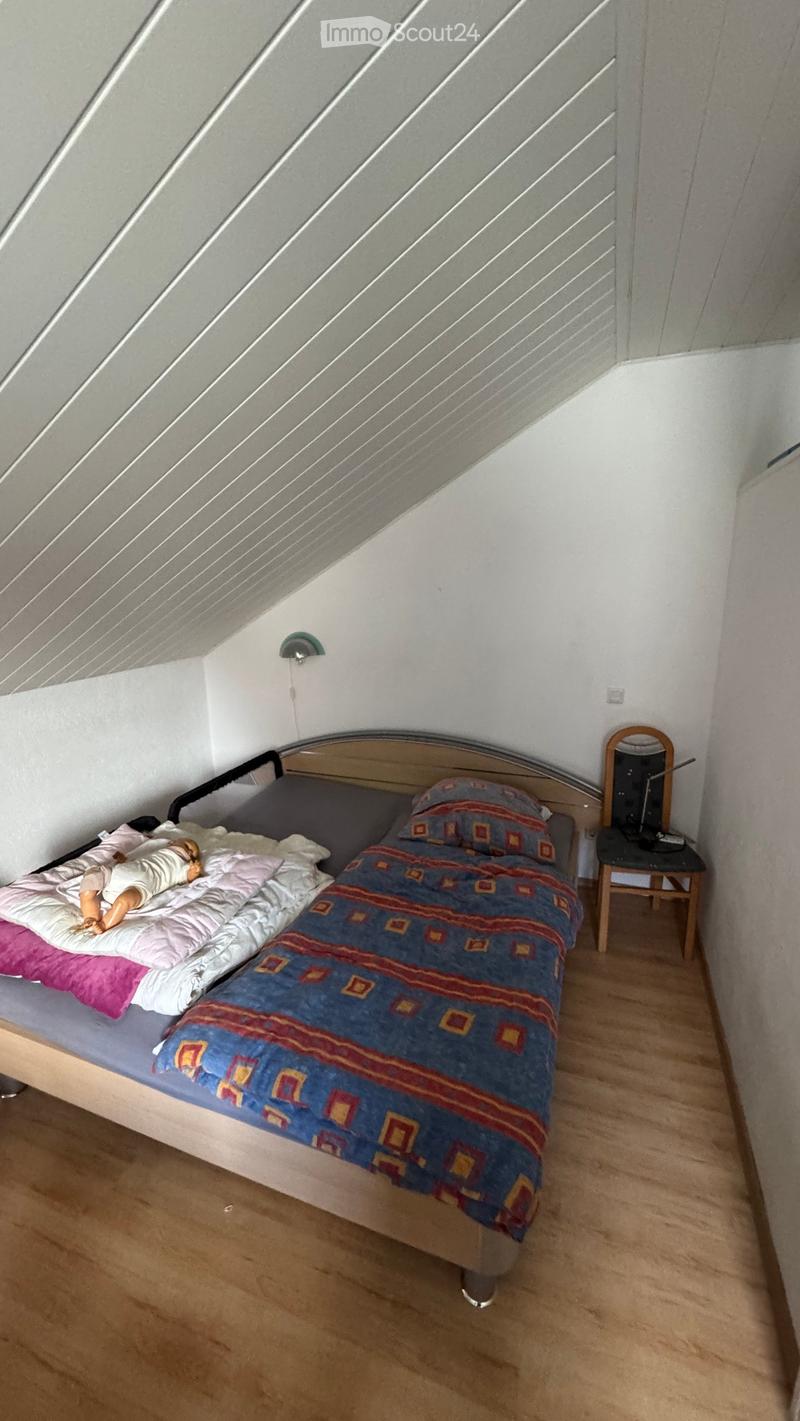 Cozy attic bedroom with wooden floor, slanted ceiling, single bed with colorful bedding, small mirror on a stool, and wall-mounted light fixture.