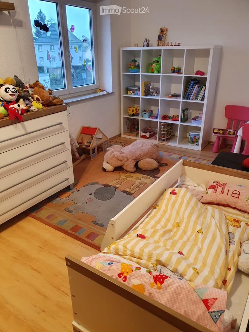 A well-lit children's bedroom with a bunk bed, colorful bedding, a large window with stuffed animals on the windowsill, a white shelving unit filled with toys and books, and a small play table.