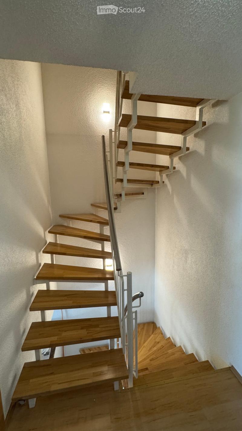 Spiral staircase with wooden steps and metal railing, illuminated by wall-mounted lights, leading to an upper level.