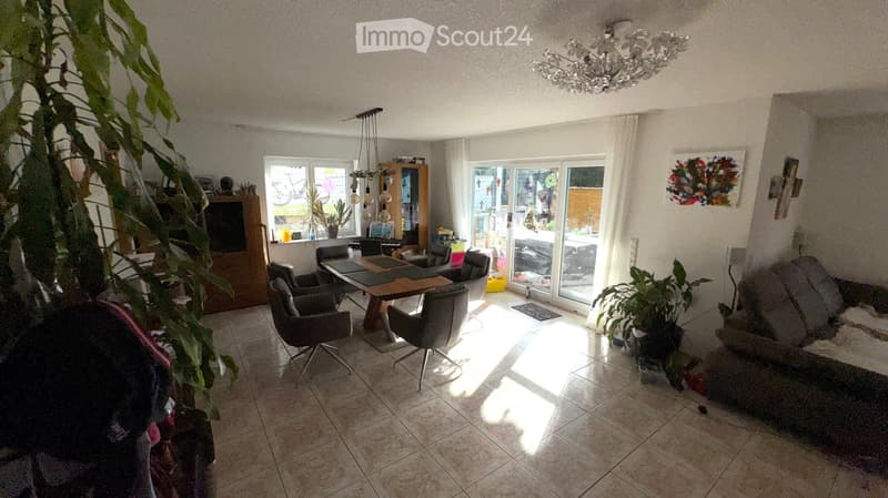 Bright living room with a sofa, dining table, chairs, chandelier, and plants. Large windows and sliding glass doors lead to an outdoor area.