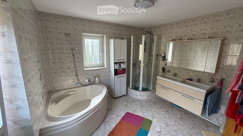 Modern bathroom with a large bathtub, double sink, mirrored cabinet, and a walk-in shower. The space features tiled walls and floor, a window for natural light, and colorful towels on the floor.