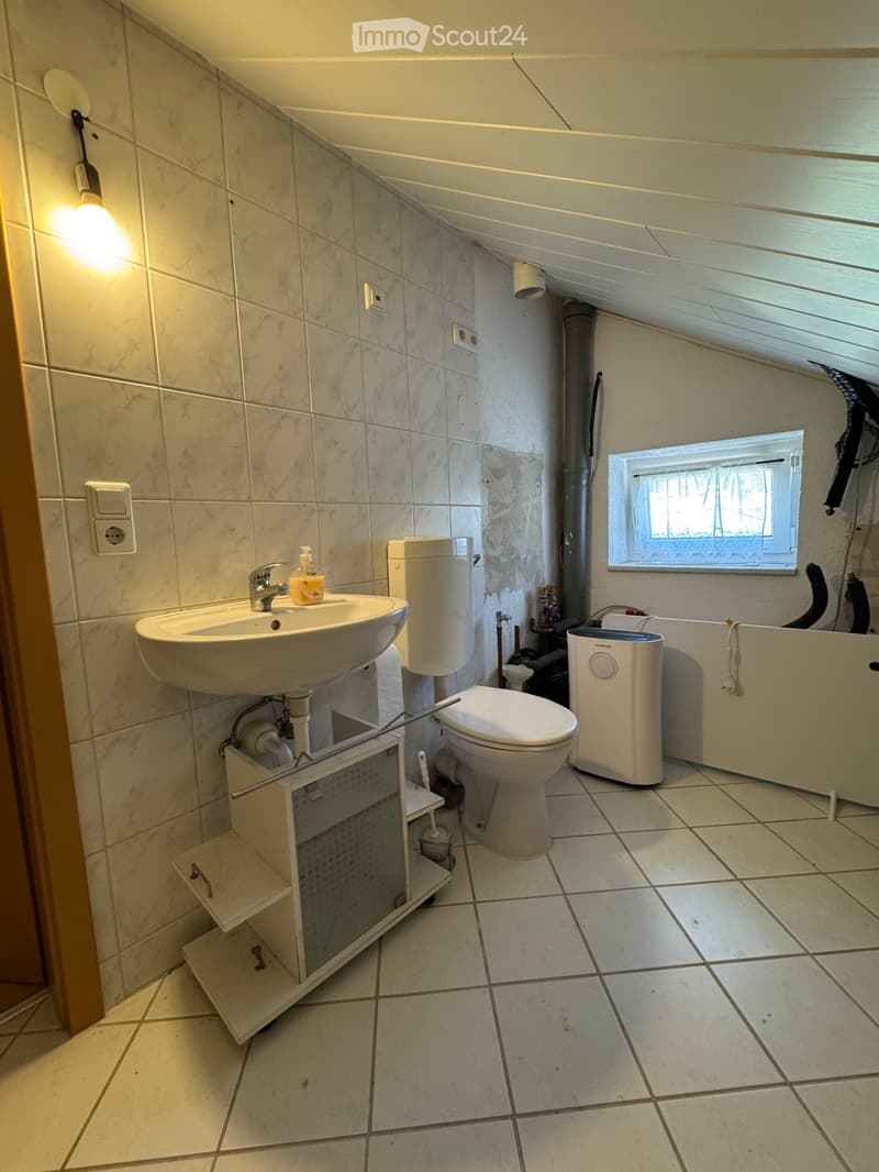 Bathroom with a sink, toilet, and a small window. The walls are tiled, and there is a light fixture on the wall.