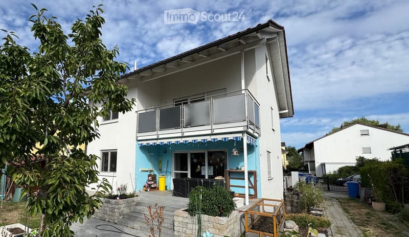 Two-story house with white exterior, balcony on the upper floor, blue storefront on the ground floor, and small garden area with plants and a tree.