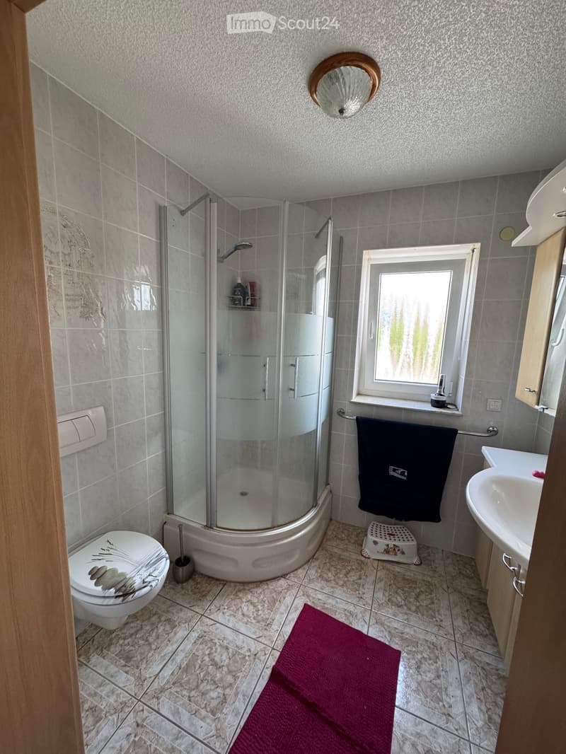 Modern bathroom with a glass-enclosed shower, toilet, sink, and a round mirror above the sink. The room features tiled walls and floor, a towel on a rack, and a small red bath mat on the floor.