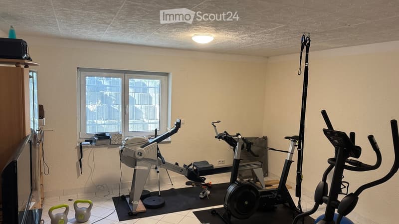 A well-lit room with beige walls and a tiled floor, featuring an elliptical trainer, treadmill, and kettlebells.