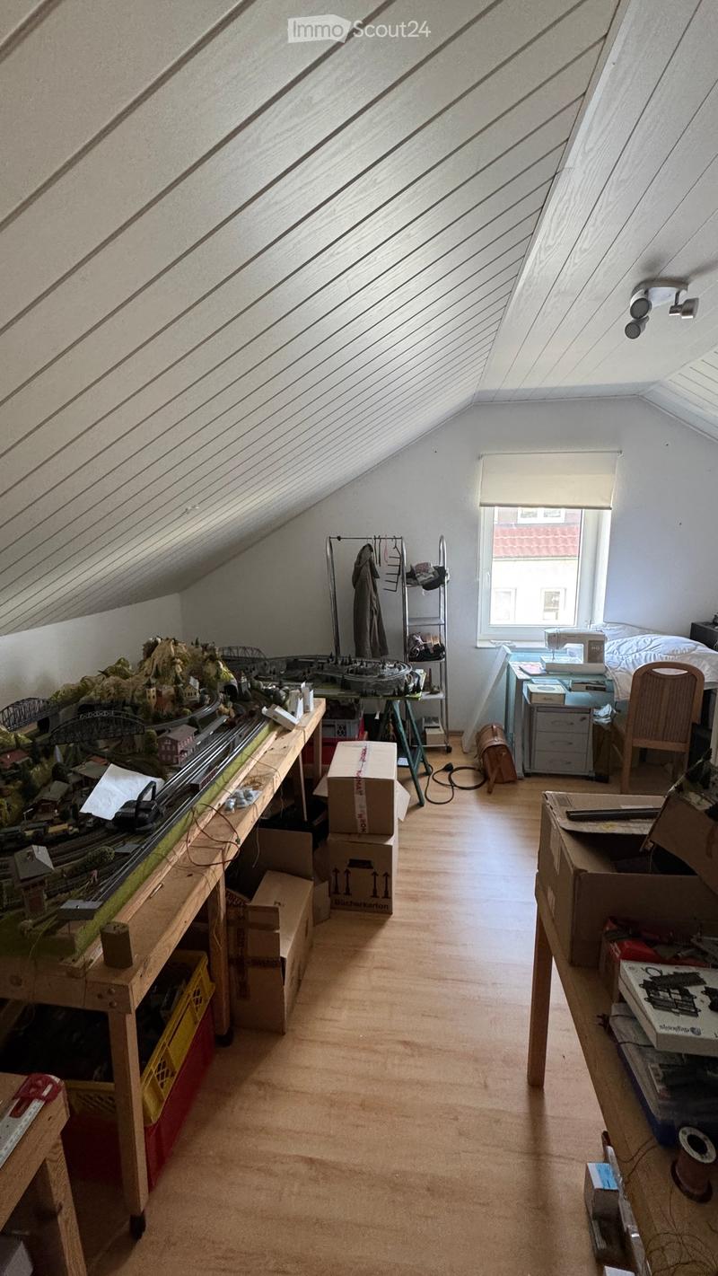 An attic room with slanted ceilings, wooden floors, and large windows. Contains a model train set on a table, a bed, a chair, and several cardboard boxes.
