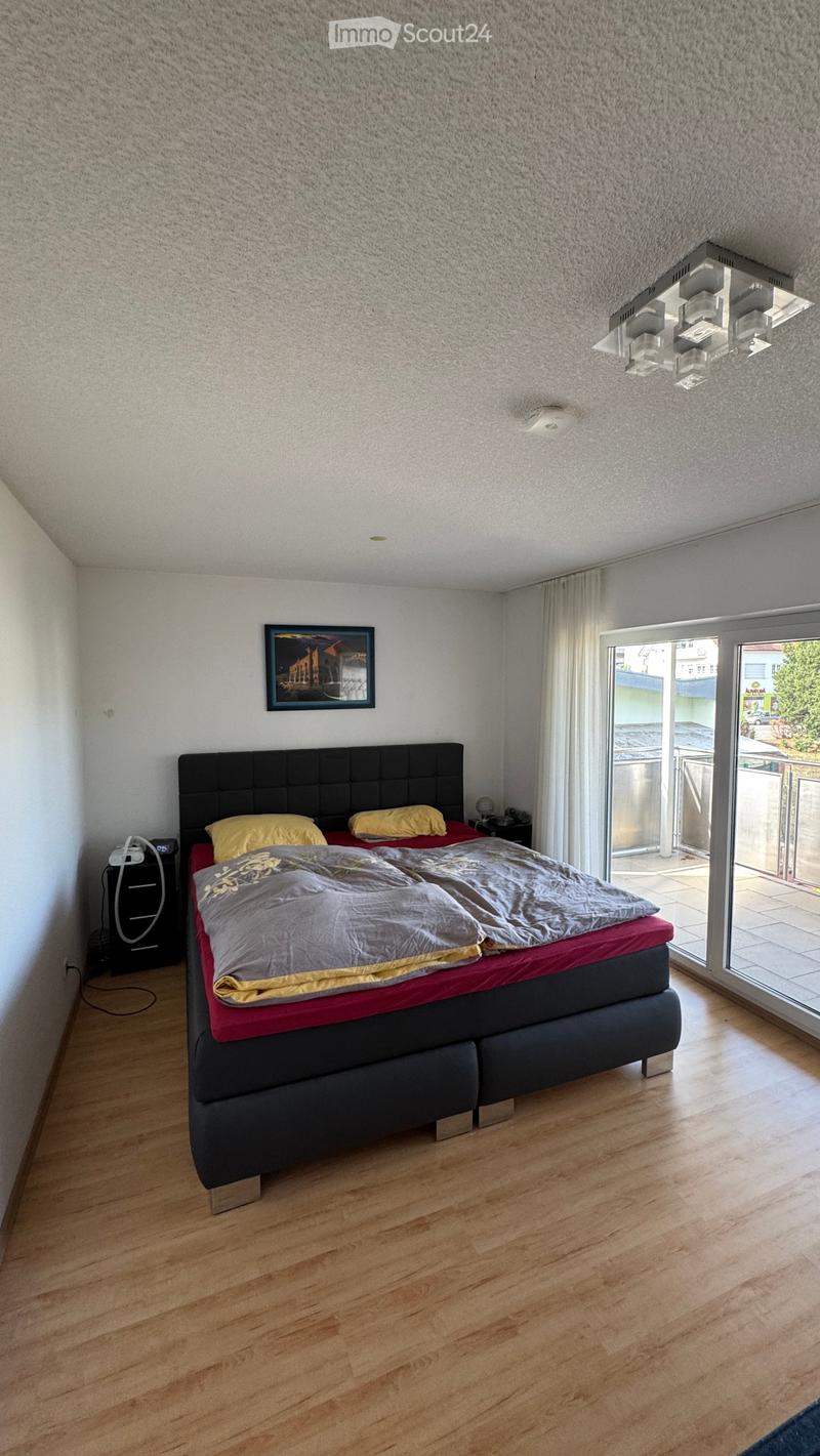 Modern bedroom with wooden flooring, large bed with dark headboard, nightstand, picture frame, and large window with curtain leading to a balcony.