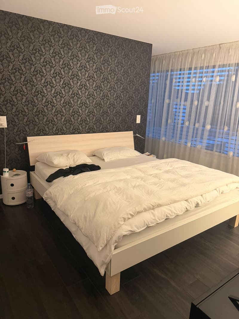 Modern bedroom with a white bed, nightstand, and dark patterned wallpaper on one wall. The bed is neatly made with white bedding and pillows, and a water bottle is placed on the floor near the nightstand.