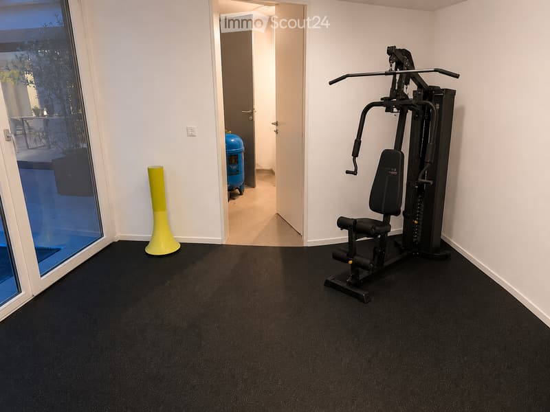 A well-lit room with a black fitness machine and a bright yellow stool, featuring a large window with a view of an outdoor area.