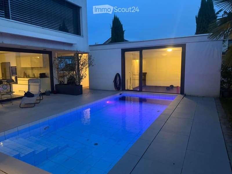 Modern outdoor area with a swimming pool, lounge chairs, and a view of the house interior through large glass doors.