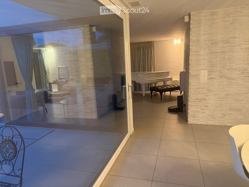 A modern, well-lit room with a white piano, a black piano bench, a sofa, a small table, and a mirror. The room features large glass doors leading to a tiled patio area.
