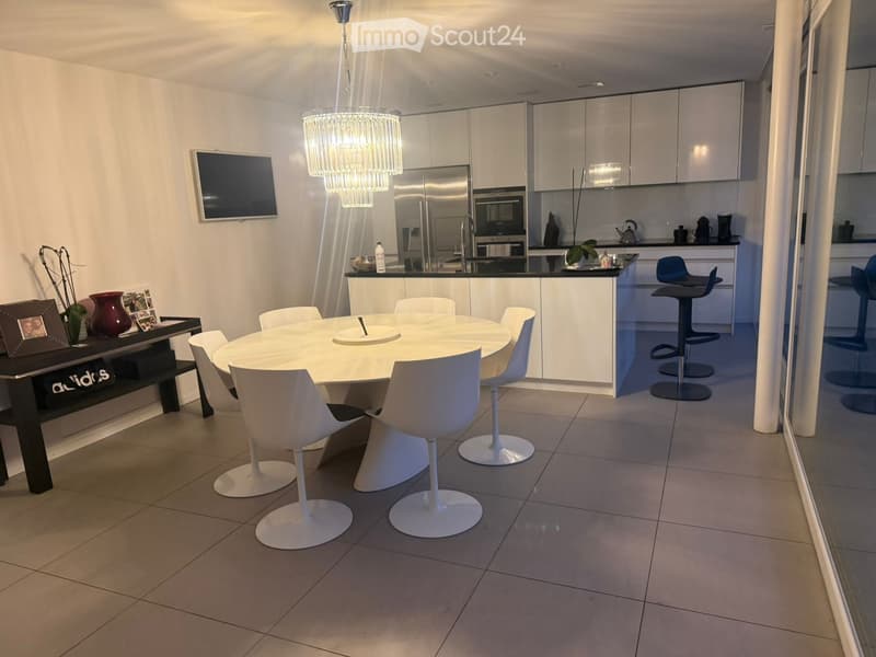 Modern open-plan dining and kitchen area with a round white dining table, white chairs, bar stools, stainless steel appliances, and a chandelier.