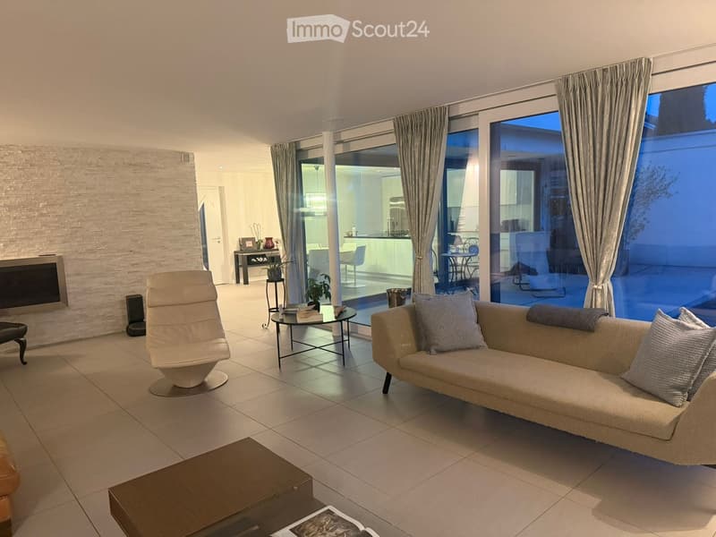 Modern living room with a beige sofa, coffee table, armchair, fireplace, and large windows offering a view of the outdoor area.