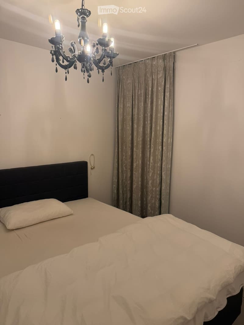 A neatly made double bed with white bedding and a black headboard, a chandelier hanging from the ceiling, and patterned curtains on the window.